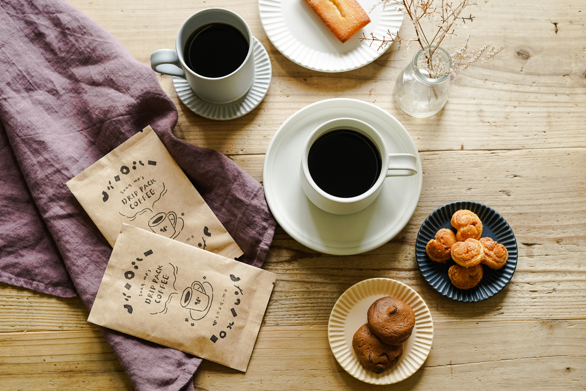 スナックミーのコーヒーパッケージ、コーヒーカップ、スナックミーの焼き菓子が盛られた皿などが並ぶテーブル
